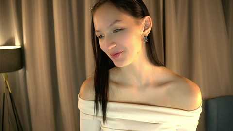 Video of an Asian woman with long black hair, wearing a beige off-shoulder top, smiling softly, in a dimly lit room with beige curtains.