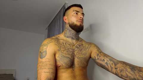 Video of a muscular, shirtless man with intricate, colorful tattoos covering his chest, arms, and neck, standing in a simple, white-walled room with a window and curtain.