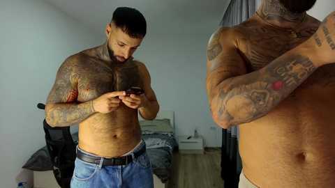 Video of a muscular, tattooed man with short dark hair, wearing jeans, standing shirtless in a bedroom, focused on his smartphone.