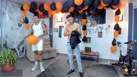 Video of two shirtless men in Halloween-themed room with spider web, orange balloons, and spider decorations; one wears ripped jeans, the other white shorts.