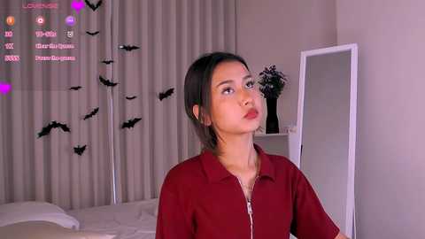Video of a young Asian woman with medium-length black hair, wearing a red blouse, looking introspective in a bedroom with white curtains and a mirror.