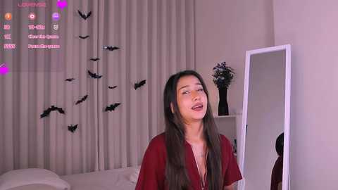 Video of an East Asian woman with long black hair, wearing a maroon top, smiling in a bedroom with white curtains, bat decorations, and a mirror.