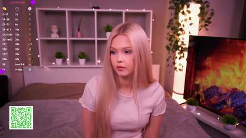 Video of a blonde young woman with fair skin and a slender build, wearing a white shirt, sitting on a bed in a cozy, dimly lit room with a TV, fireplace, and plants.