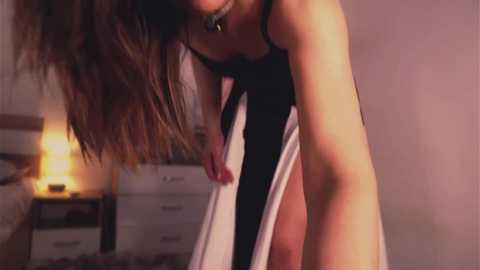 A video of a slender, brunette woman in a black tank top, pulling up white panties, in a dimly lit bedroom with a white dresser and warm, glowing lamp.