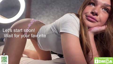 Video of a light-skinned woman with long brown hair, wearing a white top and pink lace panties, kneeling on a bed, smiling seductively. Text reads, \"Let's start soon! Wait for your favorite.\