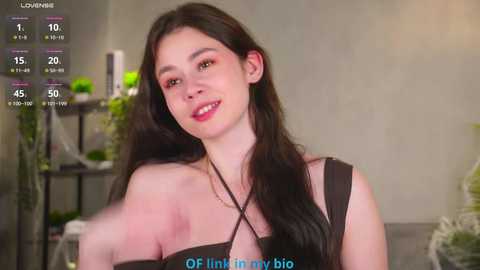 Video of a young woman with long black hair, wearing a black off-shoulder top, smiling. Background includes a green plant and a shelf with vases. Text reads \"LOVINNI,\" \"OF LINK TO MY BIO,\" and \"45.\