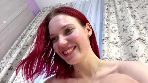 Media: Video of a smiling, fair-skinned woman with red hair, topless, standing in a bathroom with white tiled walls and floral-patterned curtains.