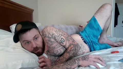 Media: Video of a tattooed man with a beard, wearing a black cap and blue shorts, lying on a bed with white sheets, in a bedroom.