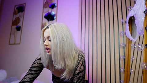 Media: Video of a blonde woman in a black, patterned sweater, bending forward with a playful expression. Background features a modern, beige and purple room with vertical blinds, decorative artwork, and a fluffy white pillow.