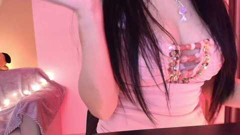 Video of a young woman with long black hair, wearing a revealing white top with a beaded design, standing in a dimly lit room with a pinkish hue.