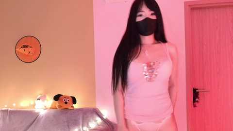 Video of an East Asian woman with long black hair, wearing a white tank top, white panties, and a black face mask, standing in a dimly lit room with a door and a wall clock.