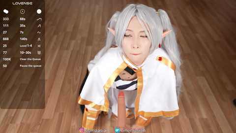 A screenshot from a video game shows an anime-style character with elf ears, twin-tailed white hair, and a white and gold cape. The character, kneeling on a wooden floor, is holding a large, erect penis.