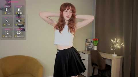Video of a slender, fair-skinned woman with long, wavy auburn hair, wearing a white crop top and black skirt, standing confidently in a modern room with a calendar, chair, and vase.