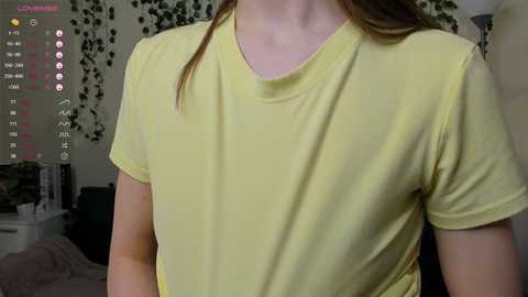 Video of a light-skinned woman with brown hair wearing a yellow T-shirt, indoors with a green leafy wall background and a TV screen showing streaming app interface.