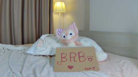 Video of a plush pink cat with a purple flower on its head, lying on a bed with white sheets and a beige pillow, next to a handwritten \"BB\" sign and a lit table lamp.