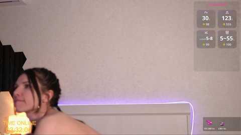 Video of a woman with brown hair, nude, facing away, in a bedroom with a white headboard and a digital clock displaying 5:55.