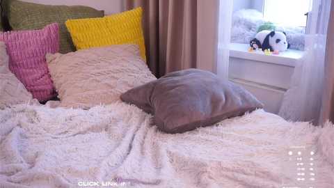 Video of a cozy bedroom with white fluffy bedding, two large pillows (one yellow, one green), a gray cushion, a window with white curtains, and a stuffed panda on the windowsill.