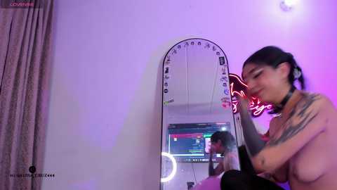A video of a topless woman with medium-sized breasts, dark hair in pigtails, wearing a choker, sitting in front of a mirror, illuminated by a ring light, in a purple-lit room with neon signage.