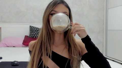 Media: Video of a young woman with long, straight, light brown hair, wearing a black off-shoulder top, sipping from a clear coffee mug, in a minimalist, white room with a bed and pink pillow.