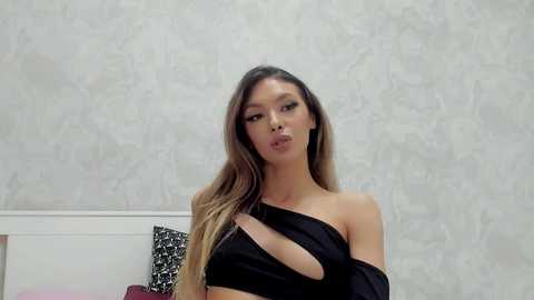 Video of a young Asian woman with long, straight, brown hair, wearing an off-shoulder black crop top, standing in a room with white wallpaper and a black and white patterned pillow.