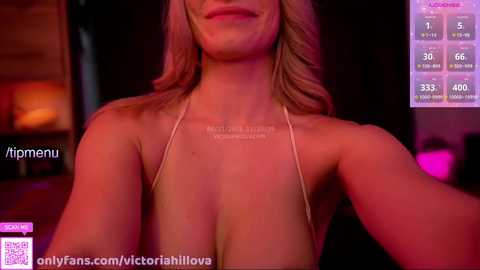 Video of a smiling, light-skinned woman with long blonde hair, wearing a revealing halter top that highlights her cleavage, taken indoors with a pinkish-purple lighting.