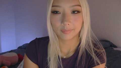Video of an East Asian woman with long, straight, platinum blonde hair, wearing a dark purple t-shirt, sitting indoors with a neutral background.