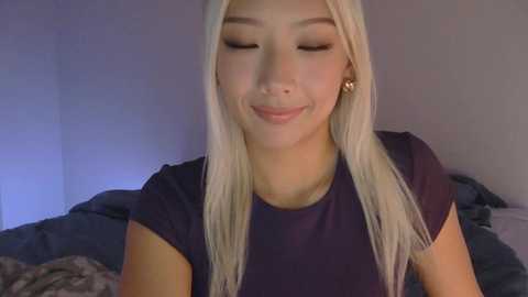 A video of a smiling Asian woman with long platinum blonde hair, wearing a dark purple top, in a softly lit bedroom.