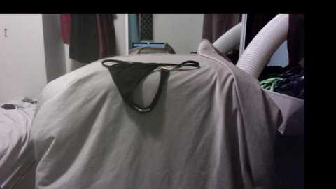A video of a messy bedroom with a white comforter and a black thong on top, beige sheets, and a beige towel draped over a white dresser.