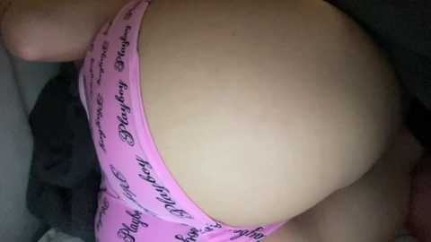 Video of a woman's lower back and buttocks, wearing pink panties with black text, lying on a bed with dark bedding, focusing on the curve of her body.