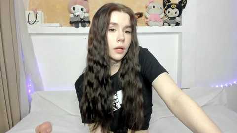 Video of a young woman with long, wavy brown hair, fair skin, and a slender build, wearing a black T-shirt. She's in a bedroom with white walls and plush toys, including Sanrio characters, in the background.