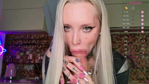 Video of a pale, platinum blonde woman with heavy makeup, wearing a black top, licking her fingers while holding a tattooed hand. Background shows a kitchen with tiled walls and a neon sign.