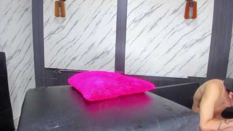 Video of a man lying shirtless on a black leather couch, with a bright pink cushion on his chest. Background features a modern interior with white marble walls and wooden accents.