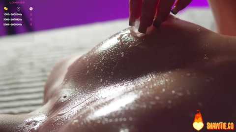 Video of a nude woman's glistening, wet back and shoulders. Her skin is dark, and she has a red manicured hand on her shoulder. Background is a blurred purple and grey.