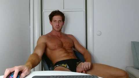Video of a shirtless, muscular man with short brown hair, wearing black briefs, sitting at a desk in a white-walled room.