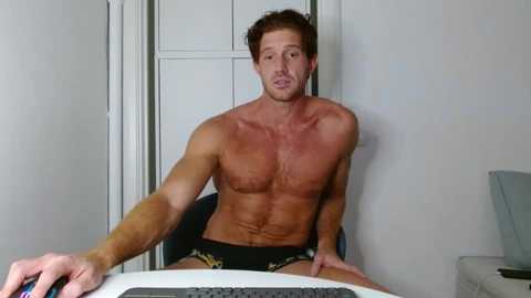 Video of a shirtless, muscular man with light skin, short brown hair, and a stern expression, wearing black boxer briefs with leopard print. He sits in front of a computer, holding a mouse. Background includes a plain white wall and part of a monitor.
