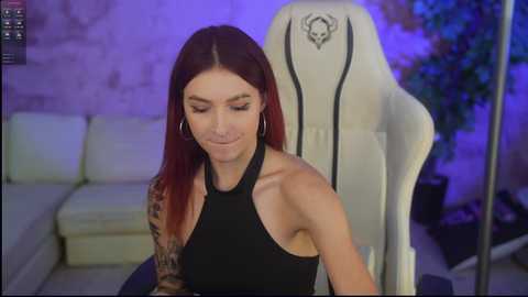 Media: Video of a tattooed woman with long, straight red hair, wearing a black halter top, sitting in a white gaming chair in a dimly lit room with a beige sofa and green potted plant.