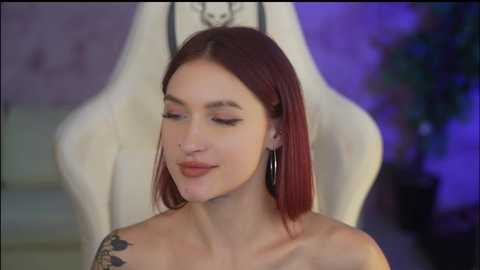 Media: Video of a fair-skinned woman with straight, shoulder-length auburn hair, wearing minimal makeup, small hoop earrings, and a tattoo on her right shoulder. She stands in front of a white gaming chair, with a purple backdrop.