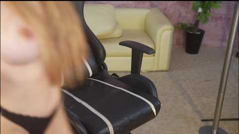 Video of a close-up of a woman's face and upper body in a black gaming chair with a yellow couch and plant in the background.
