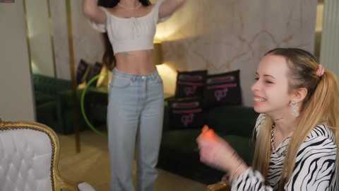 Video of two young women dancing. One, with long blonde hair in a ponytail, wears a white crop top and jeans, grinning. The other, with long dark hair, dons a white crop top and light-wash jeans. They're in a plush, dimly lit room with green couches, black pillows, and a zebra-print chair.