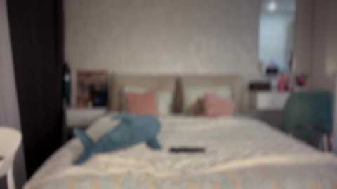 The image is a blurred video of a bedroom with a white bed, two pink pillows, a teal stuffed animal, and a black remote on the bed. The room has white walls, a window, and a white desk.