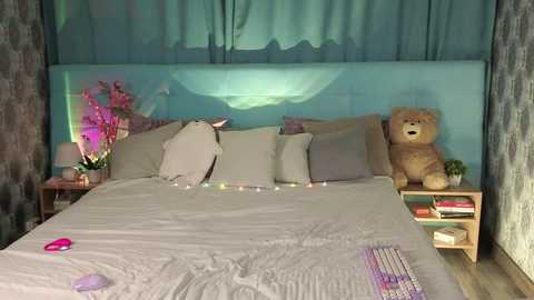 Video of a cozy, dimly-lit bedroom with a light-gray bed, teal wall art, a stuffed teddy bear, and colorful fairy lights.