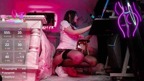 Video of a young woman with shoulder-length black hair, wearing a white top and black shorts, playing a piano in a dimly lit room with pink and purple neon lights, surrounded by gaming equipment.