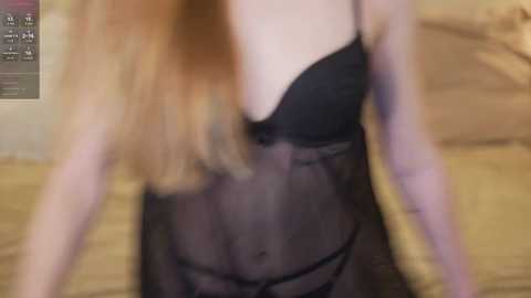 Video of a fair-skinned woman with long, wavy blonde hair, wearing a sheer black lace lingerie set, with a blurred indoor background.
