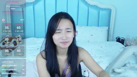 A young Asian woman with long black hair, fair skin, and a small frame smiles while sitting on a bed with white sheets and a blue upholstered headboard. The room is softly lit, with a virtual camera overlay showing her stats and a raccoon emoji.