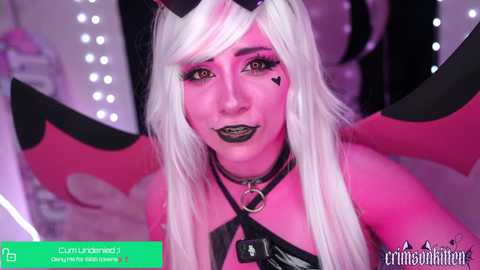 Video of a pale-skinned person with long white hair and black makeup, wearing a black choker, a black harness, and devil horns, set against a pink and black backdrop with heart-shaped cutouts.