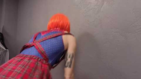 Video of a person with bright orange hair, wearing a red plaid dress, leaning against a textured gray wall. Visible tattoos on the arm.