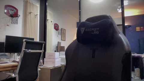 Video of a modern office desk with a black gaming chair, monitor, printer, and papers, basketball hoop on wall, white vertical blinds, and blue wall in the background.