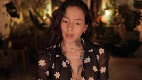 Video of a young woman with long, wavy hair, wearing a sheer black top with white flower patterns, standing indoors among green plants, tattoos on her chest and neck.