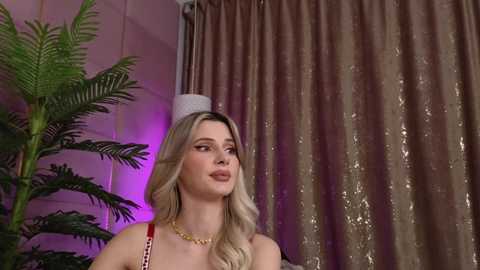 Media: Video of a blonde woman with long wavy hair, wearing a red lace bralette and a gold necklace, standing in a room with a green potted plant, beige curtains, and purple lighting.