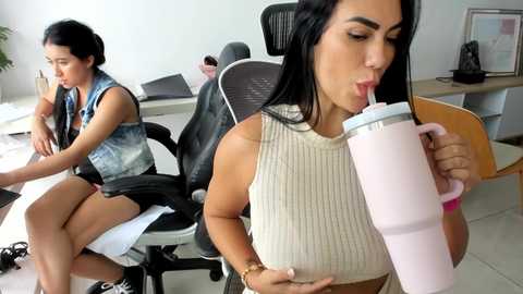 Video of two women in an office: one drinking from a pink straw, the other reading a book. They sit on black office chairs with white desks and computers in the background.